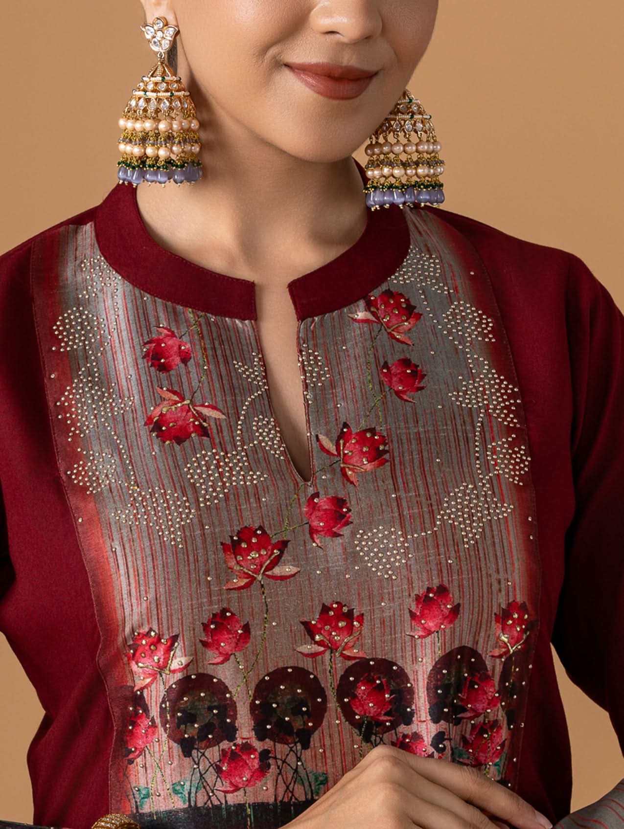 Woman wearing a maroon top with floral embroidery and gold earrings against a beige background
