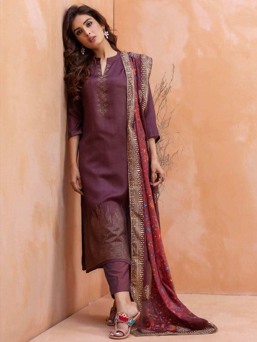 Woman wearing a traditional outfit with a purple kurta and brown pants against a beige wall.