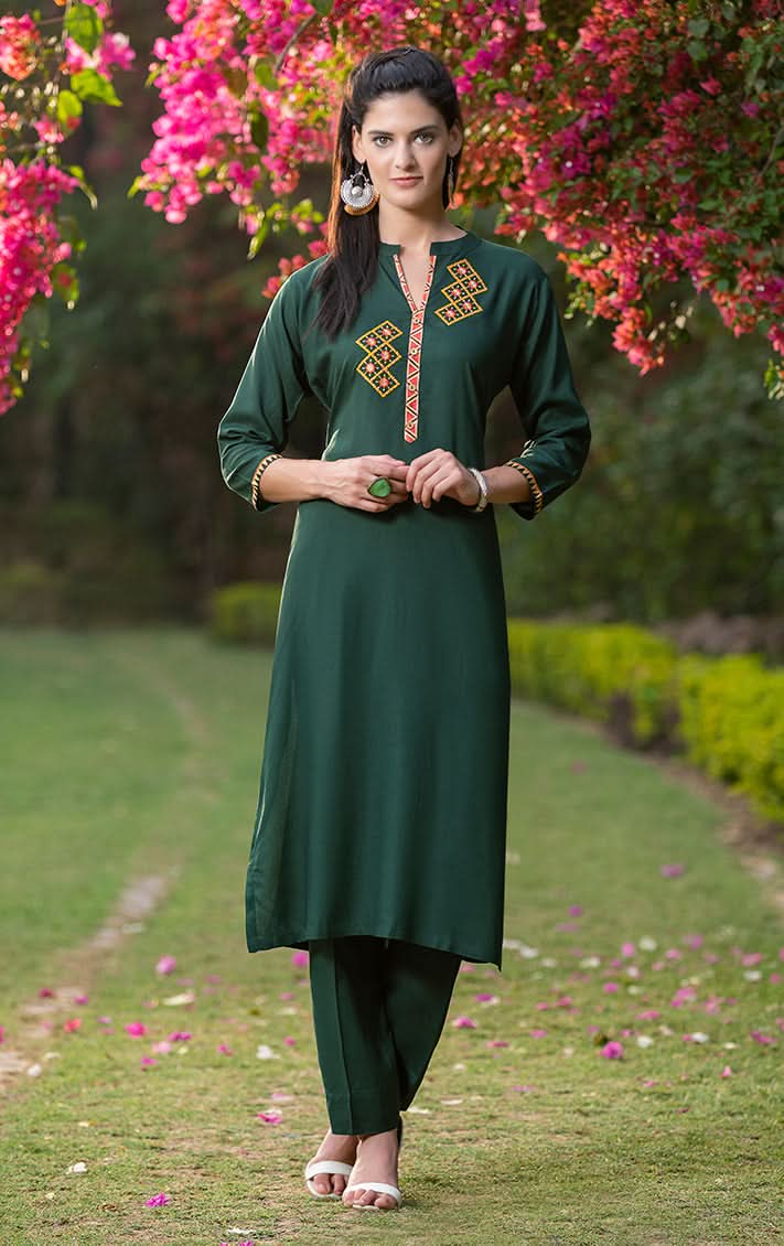 Woman in a green traditional outfit standing in a garden with pink flowers.