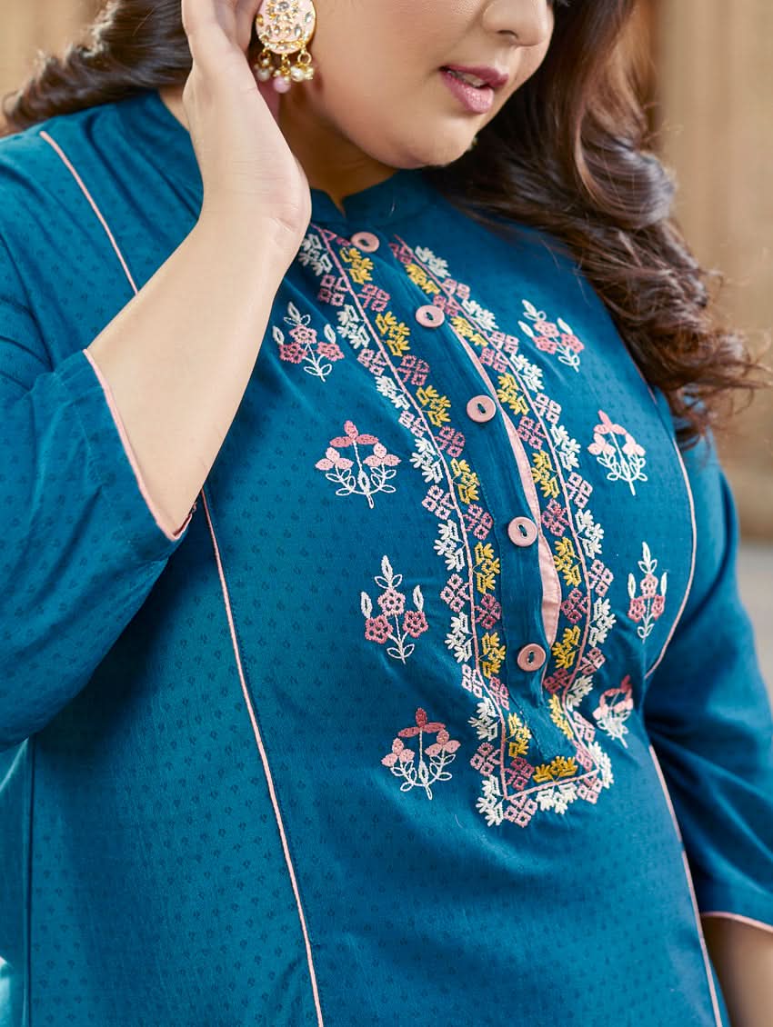 A woman standing in a beautifully decorated outdoor setting wearing a blue chunan kurta with embroidered details and three-fourth sleeves, paired with pink pants.