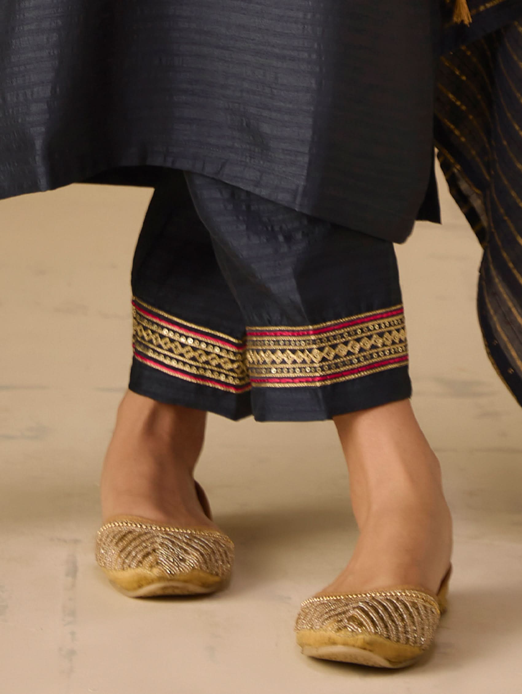 Traditional attire with navy pants featuring decorative patterns and gold sandals on a neutral background