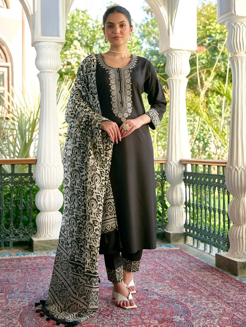 A woman standing in a columned hallway wearing a black rayon suit set with embroidered details, consisting of a kurta with round neckline and three-fourth sleeves, and pants with a partially elasticated waistband. The dupatta is also visible, laid out on the ground behind her.