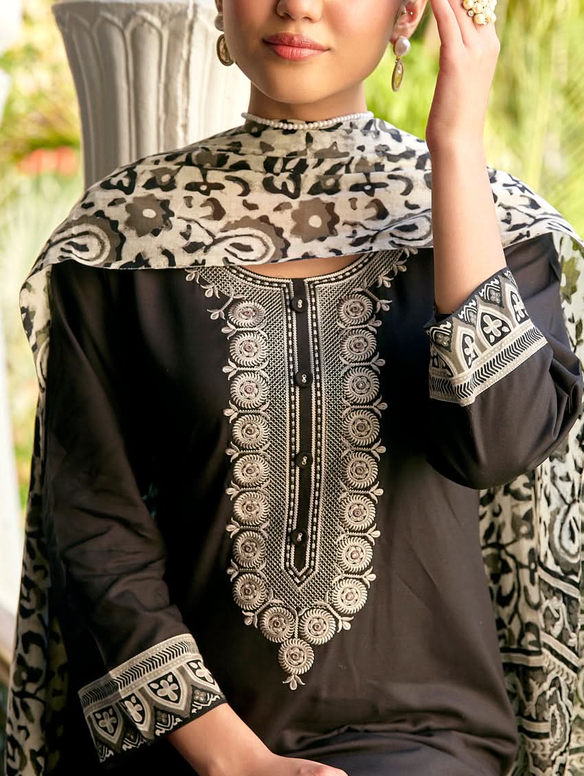 A woman standing in a columned hallway wearing a black rayon suit set with embroidered details, consisting of a kurta with round neckline and three-fourth sleeves, and pants with a partially elasticated waistband. The dupatta is also visible, laid out on the ground behind her.