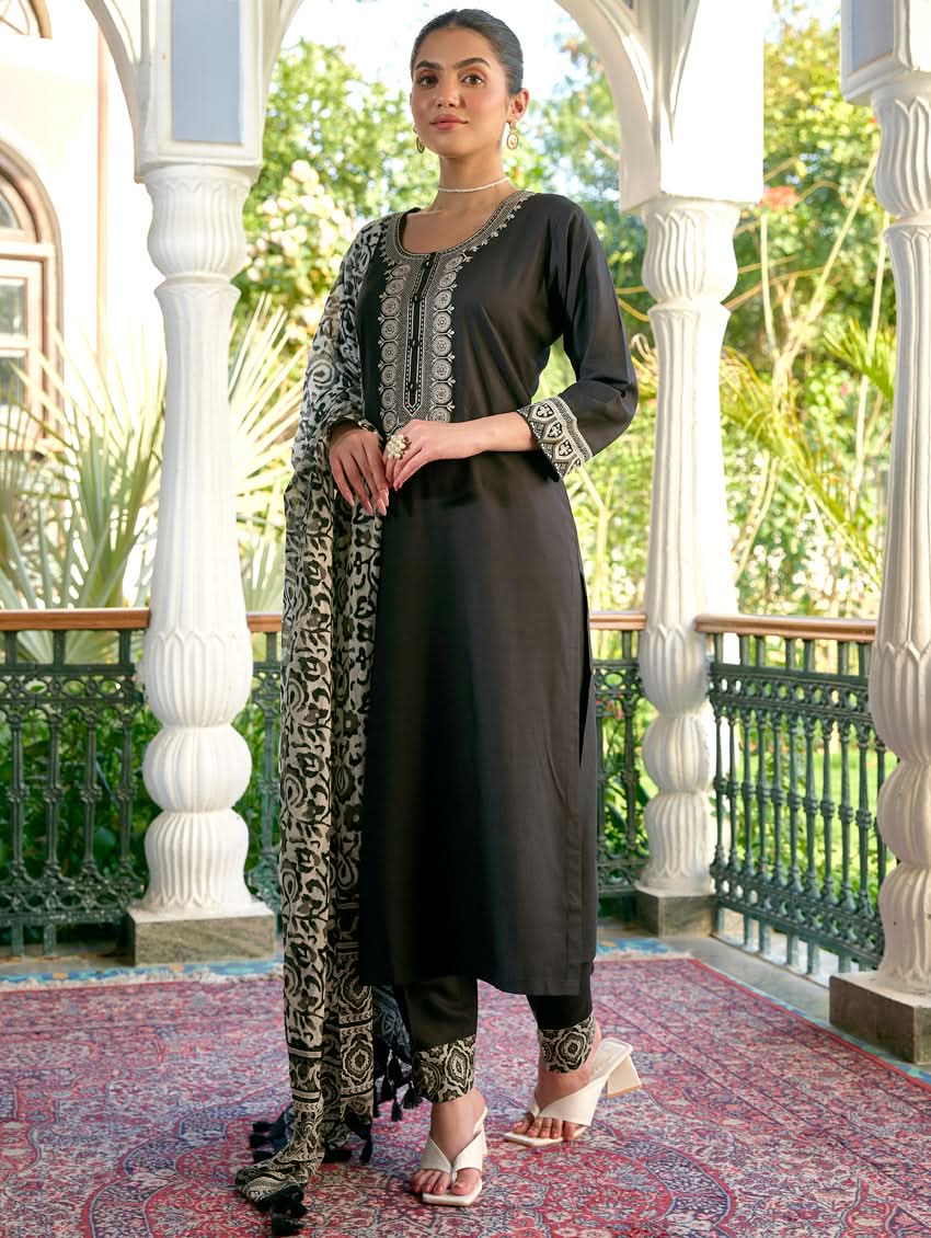 A woman standing in a columned hallway wearing a black rayon suit set with embroidered details, consisting of a kurta with round neckline and three-fourth sleeves, and pants with a partially elasticated waistband. The dupatta is also visible, laid out on the ground behind her.
