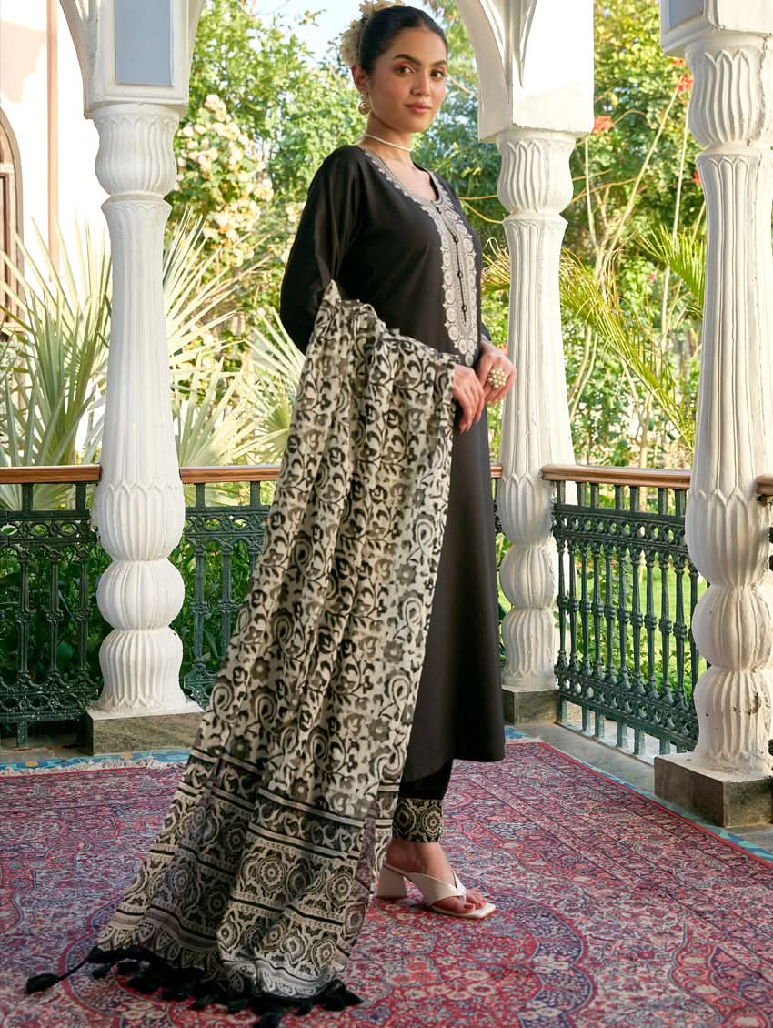 A woman standing in a columned hallway wearing a black rayon suit set with embroidered details, consisting of a kurta with round neckline and three-fourth sleeves, and pants with a partially elasticated waistband. The dupatta is also visible, laid out on the ground behind her.