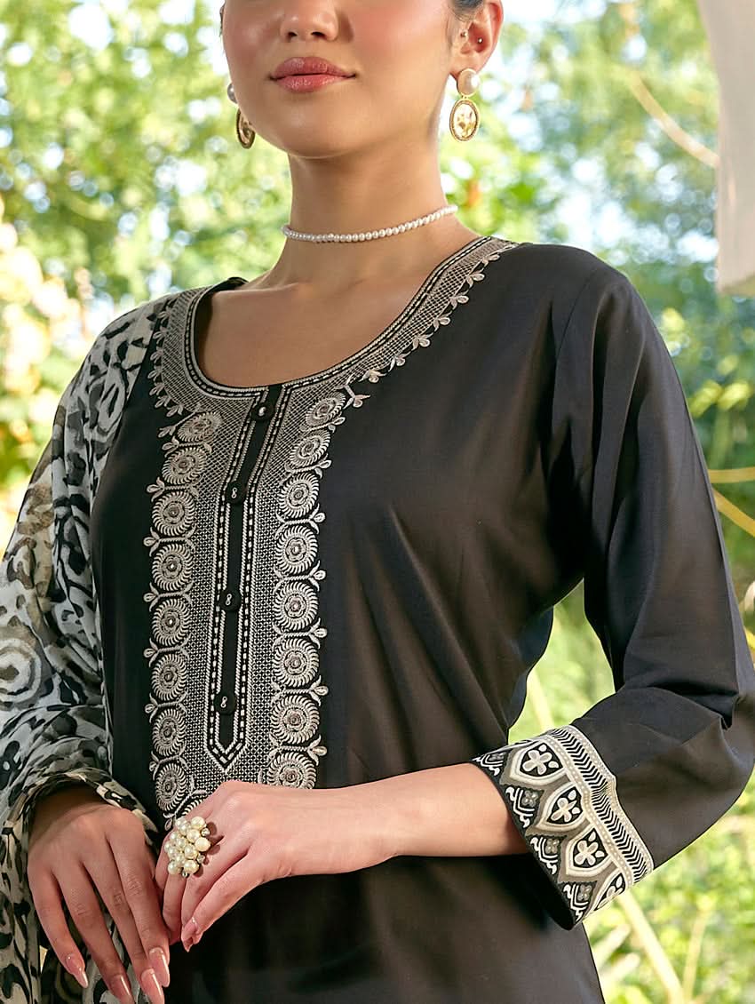 A woman standing in a columned hallway wearing a black rayon suit set with embroidered details, consisting of a kurta with round neckline and three-fourth sleeves, and pants with a partially elasticated waistband. The dupatta is also visible, laid out on the ground behind her.