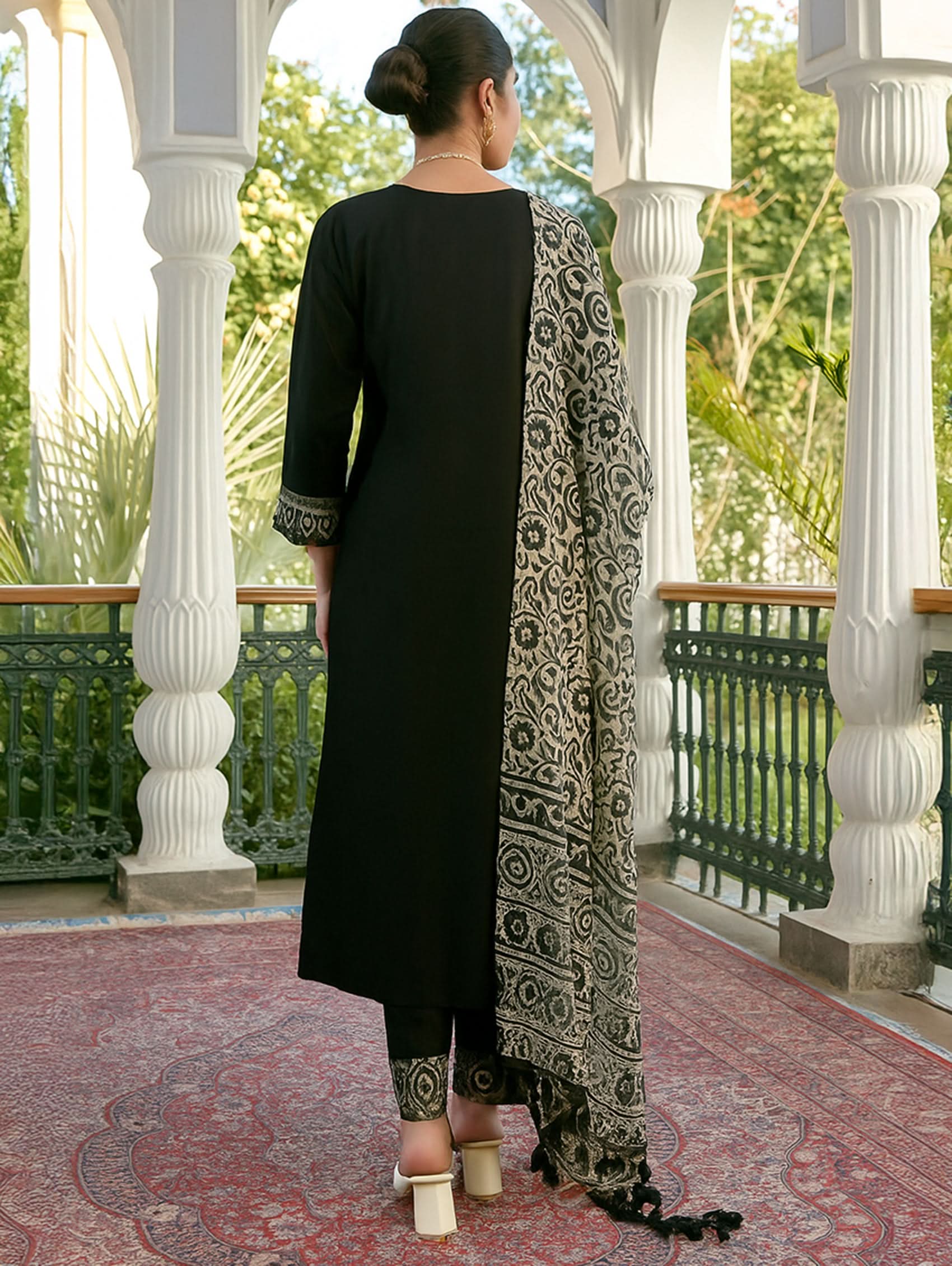 Woman in a black outfit with a patterned scarf standing on a balcony with white columns and greenery.