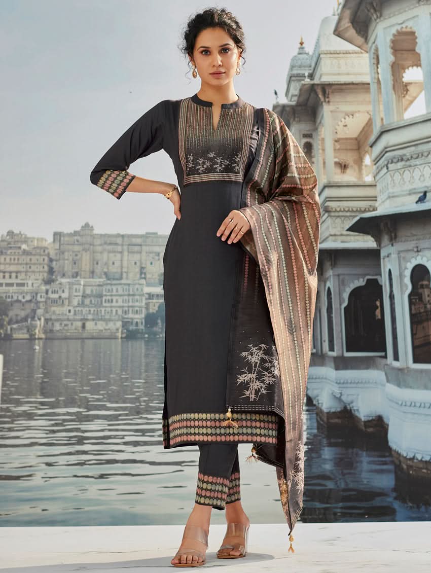 A woman wearing a traditional suit set with a kurta, pants, and a dupatta, standing near a historical building with a body of water in the background.