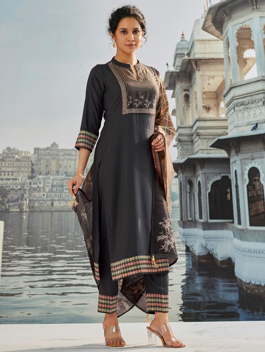 A woman wearing a traditional suit set with a kurta, pants, and a dupatta, standing near a historical building with a body of water in the background.