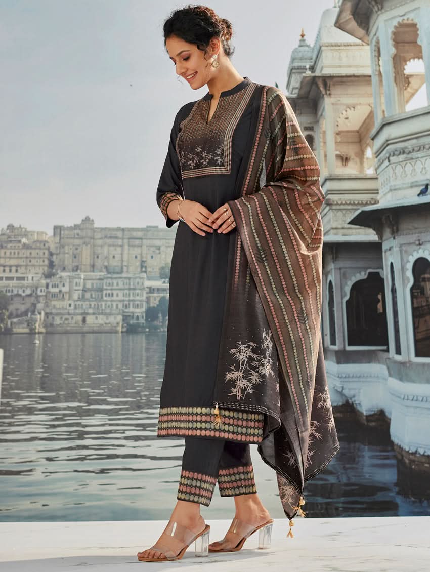 A woman wearing a traditional suit set with a kurta, pants, and a dupatta, standing near a historical building with a body of water in the background.