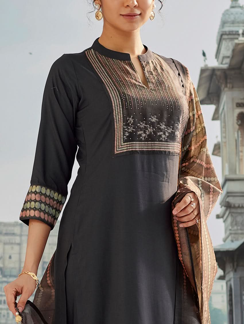 A woman wearing a traditional suit set with a kurta, pants, and a dupatta, standing near a historical building with a body of water in the background.