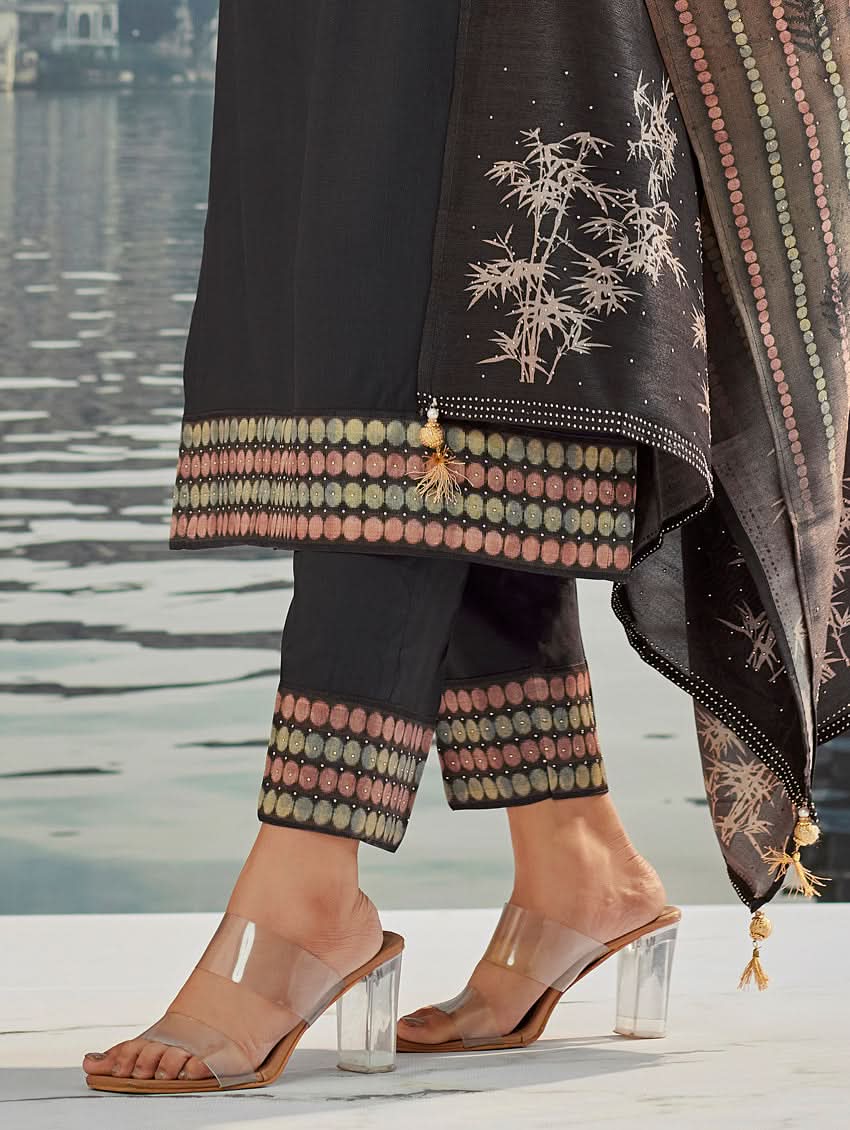 A woman wearing a traditional suit set with a kurta, pants, and a dupatta, standing near a historical building with a body of water in the background.