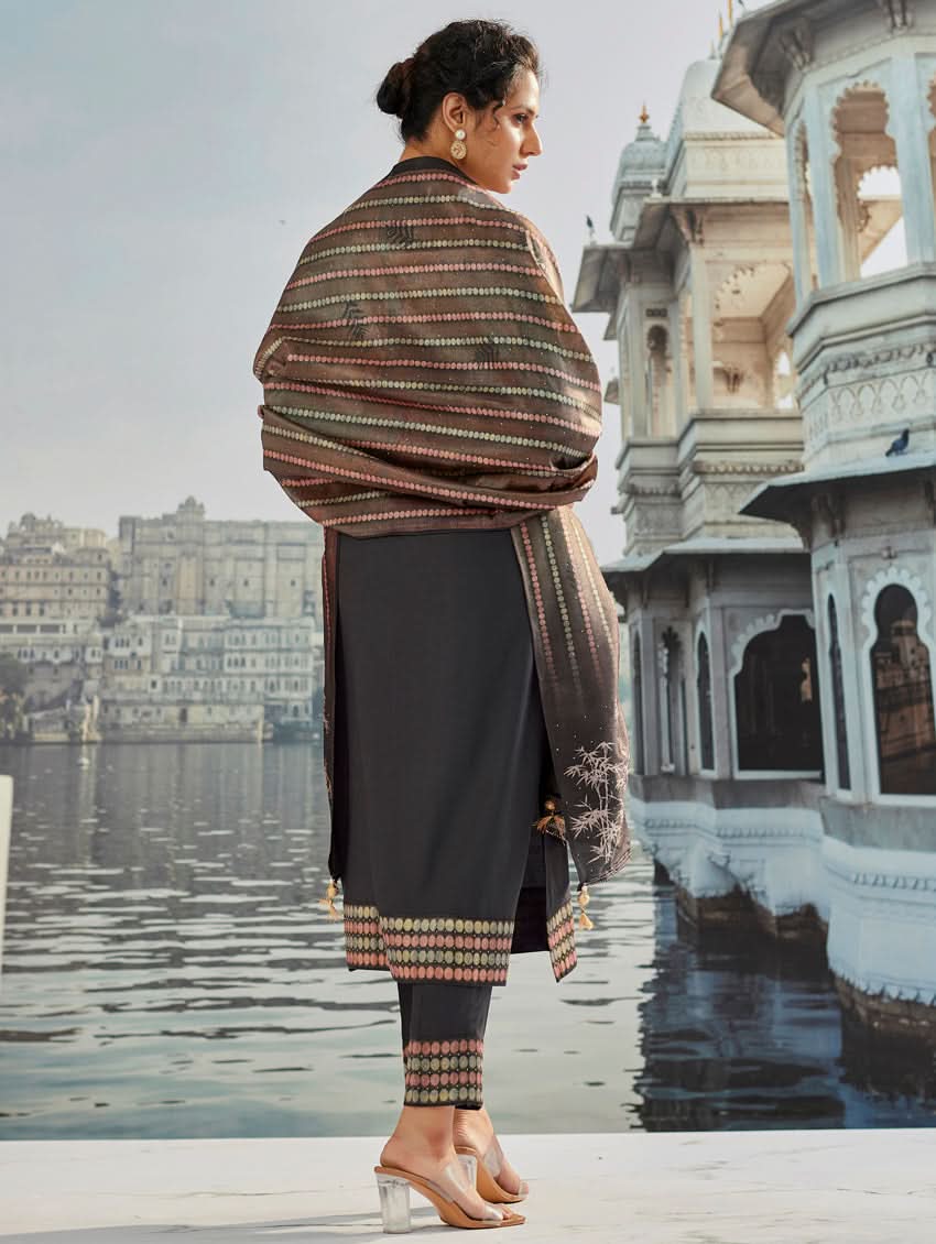 A woman wearing a traditional suit set with a kurta, pants, and a dupatta, standing near a historical building with a body of water in the background.