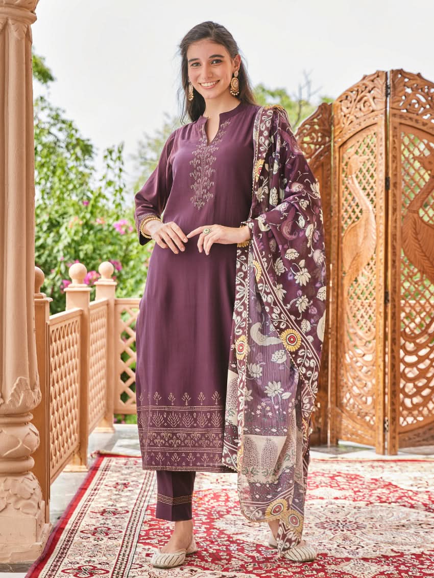 A woman standing in an elegant pose wearing a purple silk suit set with floral patterns on the dupatta, straight pants, and a kurta with pearl work.