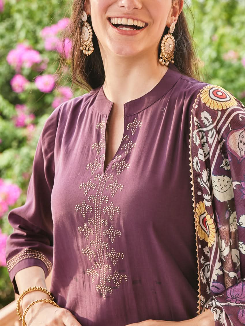 A woman standing in an elegant pose wearing a purple silk suit set with floral patterns on the dupatta, straight pants, and a kurta with pearl work.