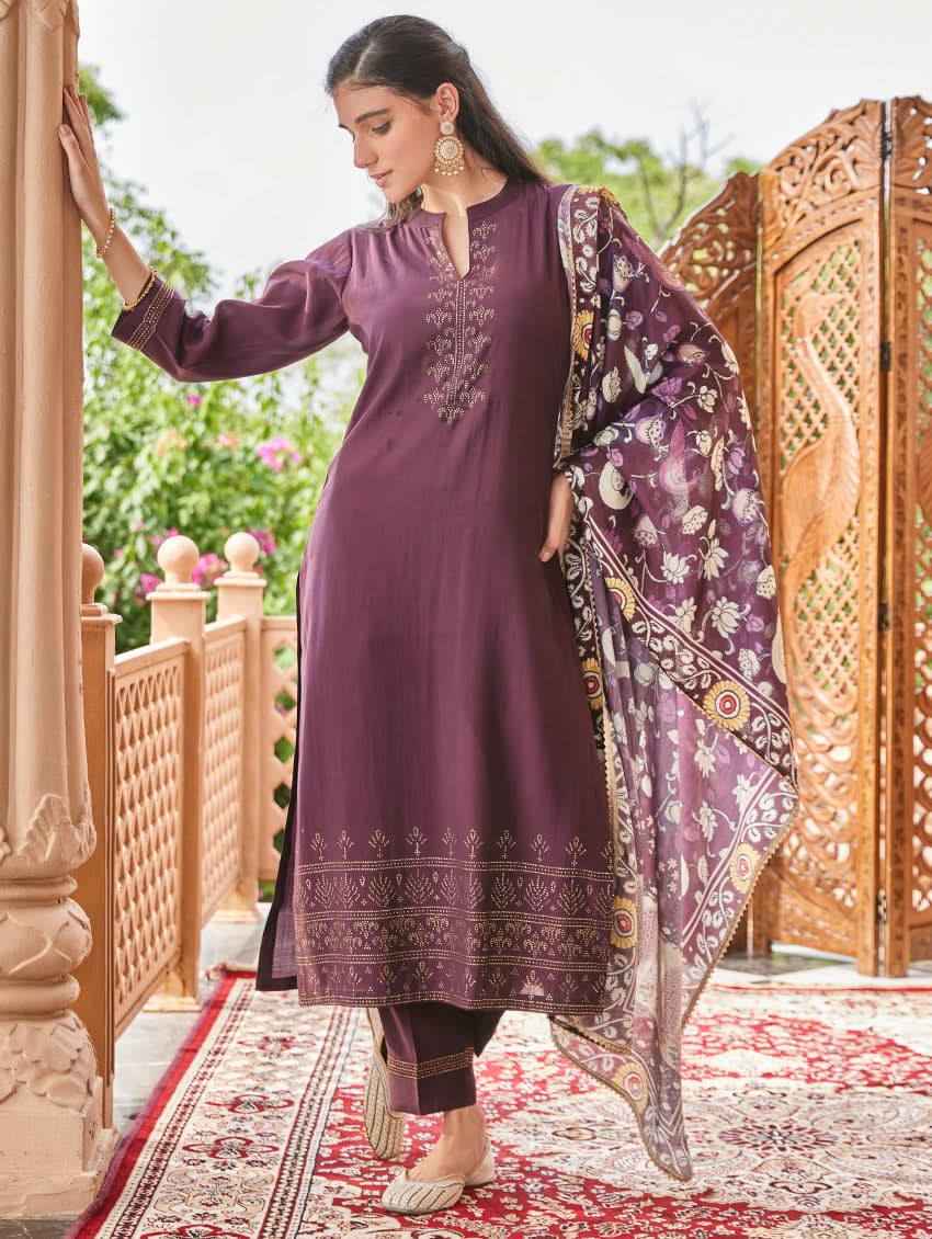 A woman standing in an elegant pose wearing a purple silk suit set with floral patterns on the dupatta, straight pants, and a kurta with pearl work.