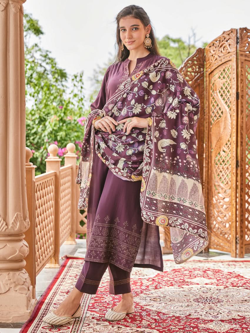 A woman standing in an elegant pose wearing a purple silk suit set with floral patterns on the dupatta, straight pants, and a kurta with pearl work.