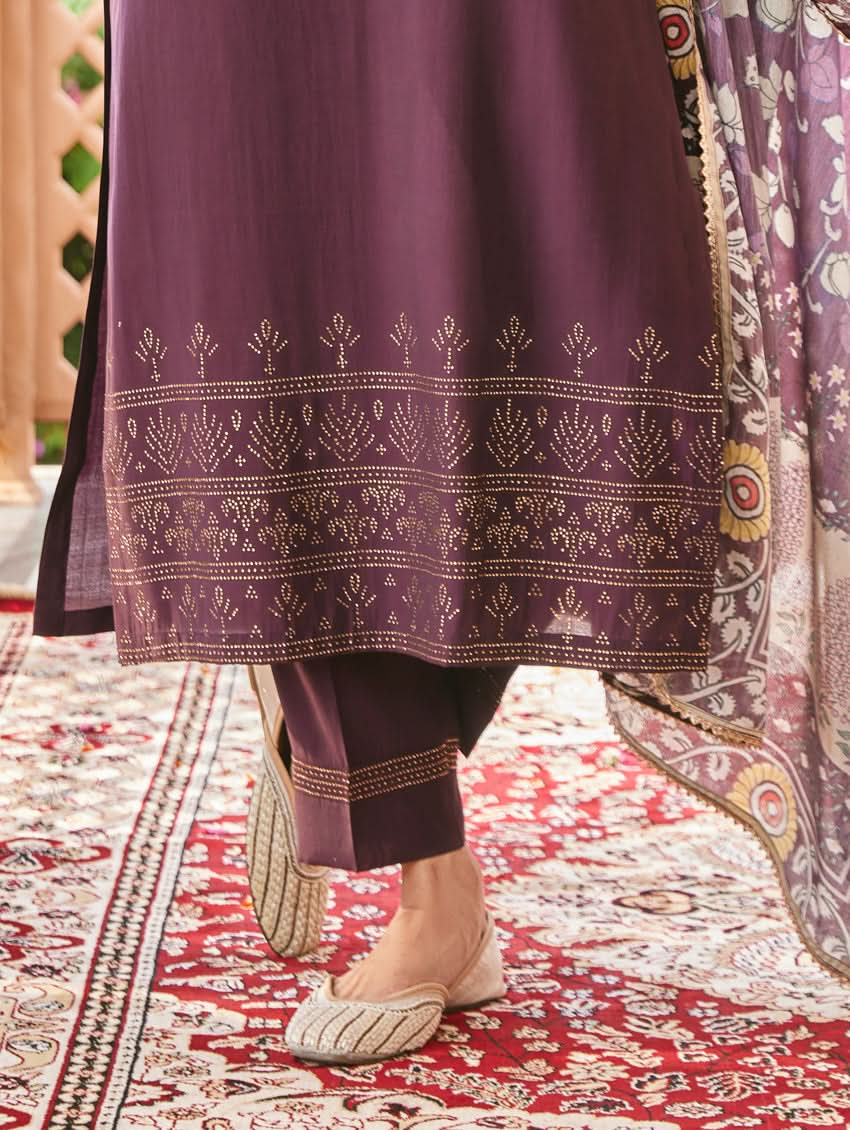A woman standing in an elegant pose wearing a purple silk suit set with floral patterns on the dupatta, straight pants, and a kurta with pearl work.