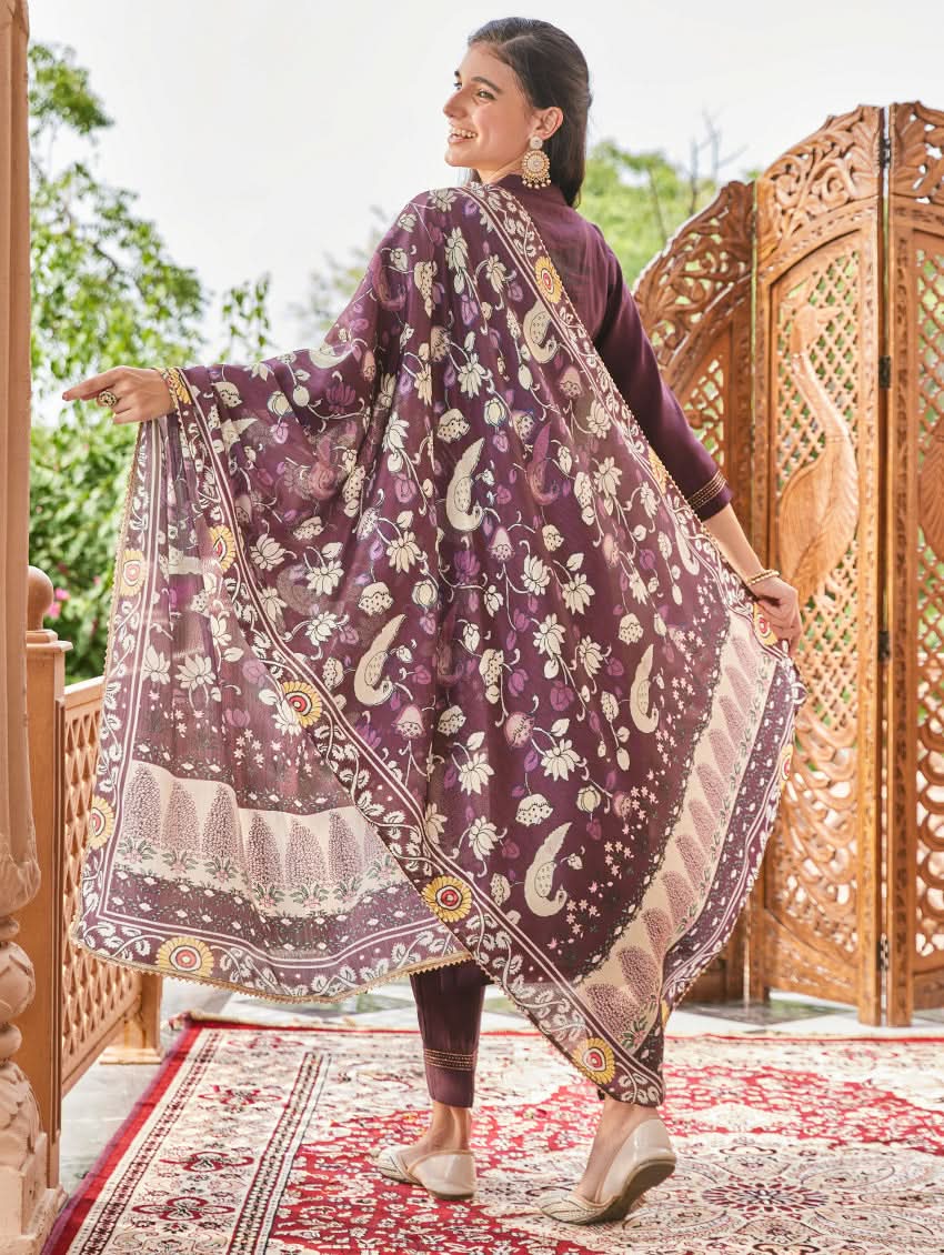 A woman standing in an elegant pose wearing a purple silk suit set with floral patterns on the dupatta, straight pants, and a kurta with pearl work.