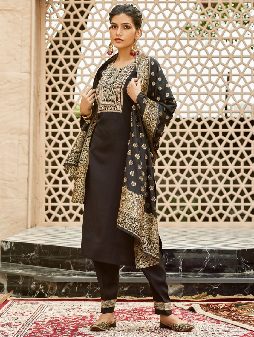 A woman wearing a traditional embroidered silk suit set with a kurta, pants, and dupatta in a Black color, standing in a decorative setting.