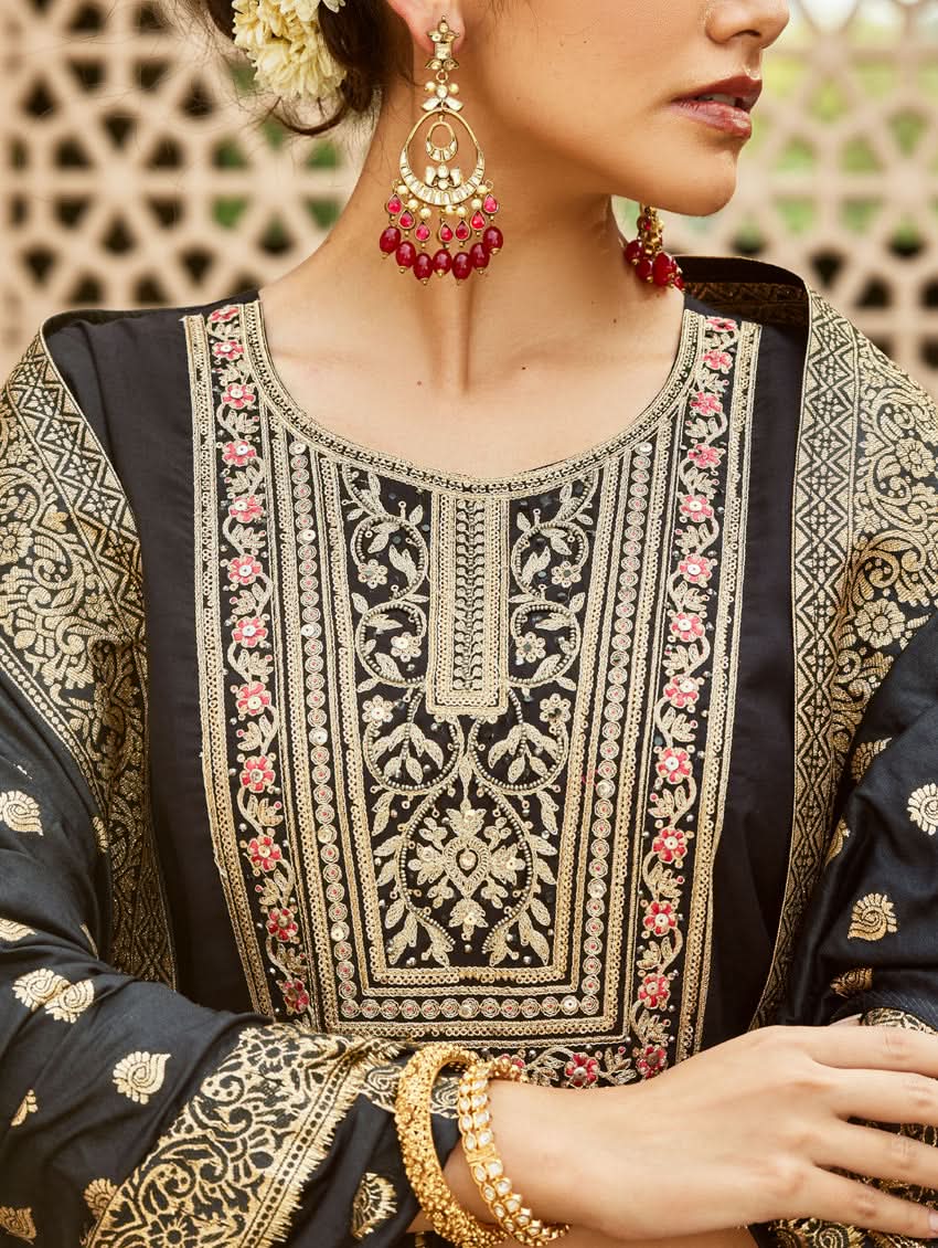 A woman wearing a traditional embroidered silk suit set with a kurta, pants, and dupatta in a Black color, standing in a decorative setting.