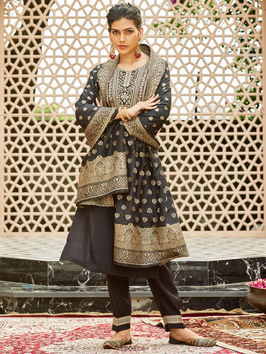 A woman wearing a traditional embroidered silk suit set with a kurta, pants, and dupatta in a Black color, standing in a decorative setting.