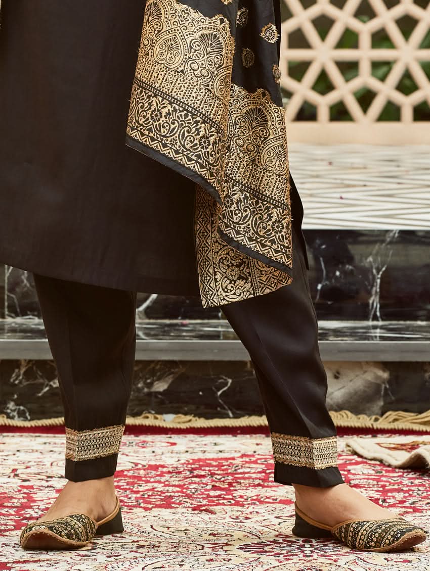 A woman wearing a traditional embroidered silk suit set with a kurta, pants, and dupatta in a Black color, standing in a decorative setting.