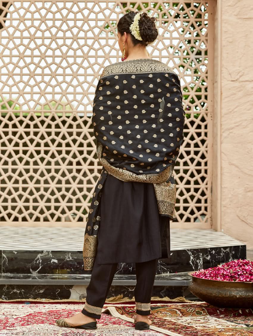 A woman wearing a traditional embroidered silk suit set with a kurta, pants, and dupatta in a Black color, standing in a decorative setting.
