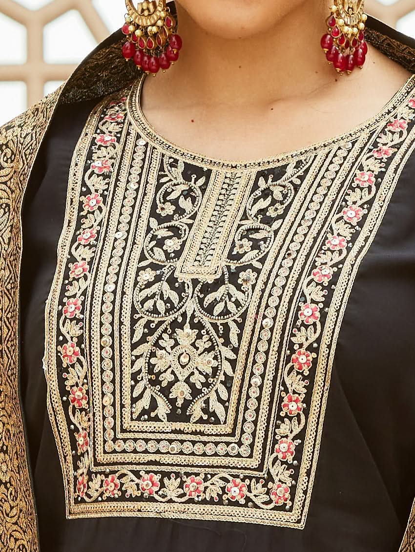 A woman wearing a traditional embroidered silk suit set with a kurta, pants, and dupatta in a Black color, standing in a decorative setting.