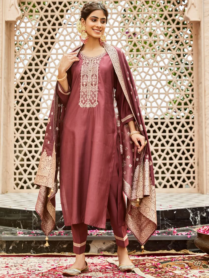 A woman wearing a traditional embroidered silk suit set with a kurta, pants, and dupatta in a purple color, standing in a decorative setting.