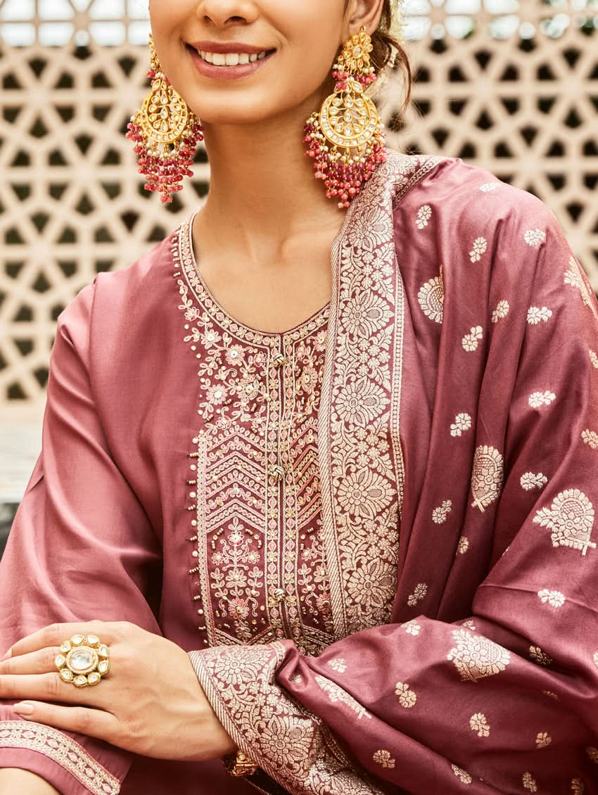 A woman wearing a traditional embroidered silk suit set with a kurta, pants, and dupatta in a purple color, standing in a decorative setting.