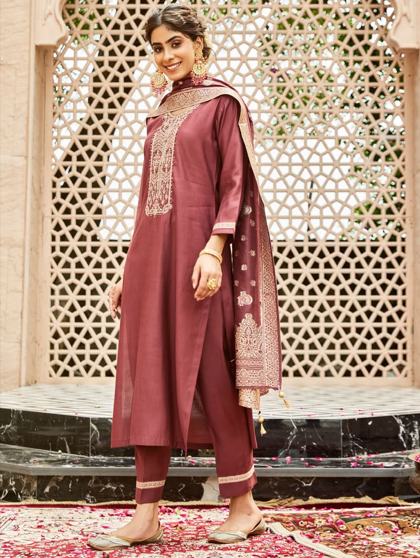 A woman wearing a traditional embroidered silk suit set with a kurta, pants, and dupatta in a purple color, standing in a decorative setting.