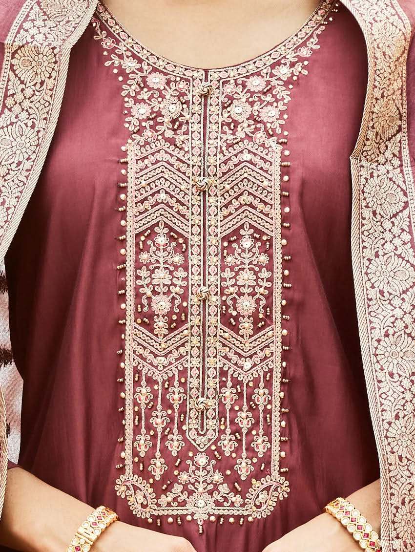 A woman wearing a traditional embroidered silk suit set with a kurta, pants, and dupatta in a purple color, standing in a decorative setting.