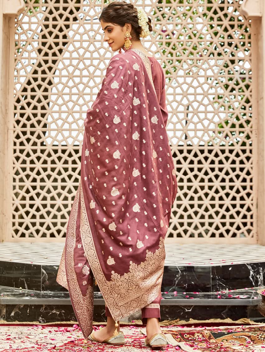 A woman wearing a traditional embroidered silk suit set with a kurta, pants, and dupatta in a purple color, standing in a decorative setting.