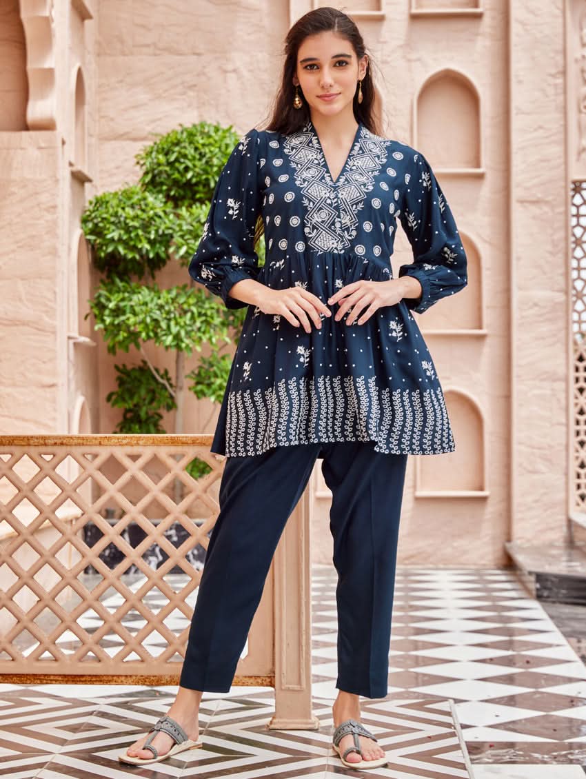 A woman standing on a balcony wearing an embroidered blue kurta set with V-neckline and three-quarter sleeves, paired with black pants with a partially elasticated waistband and pockets on either side.
