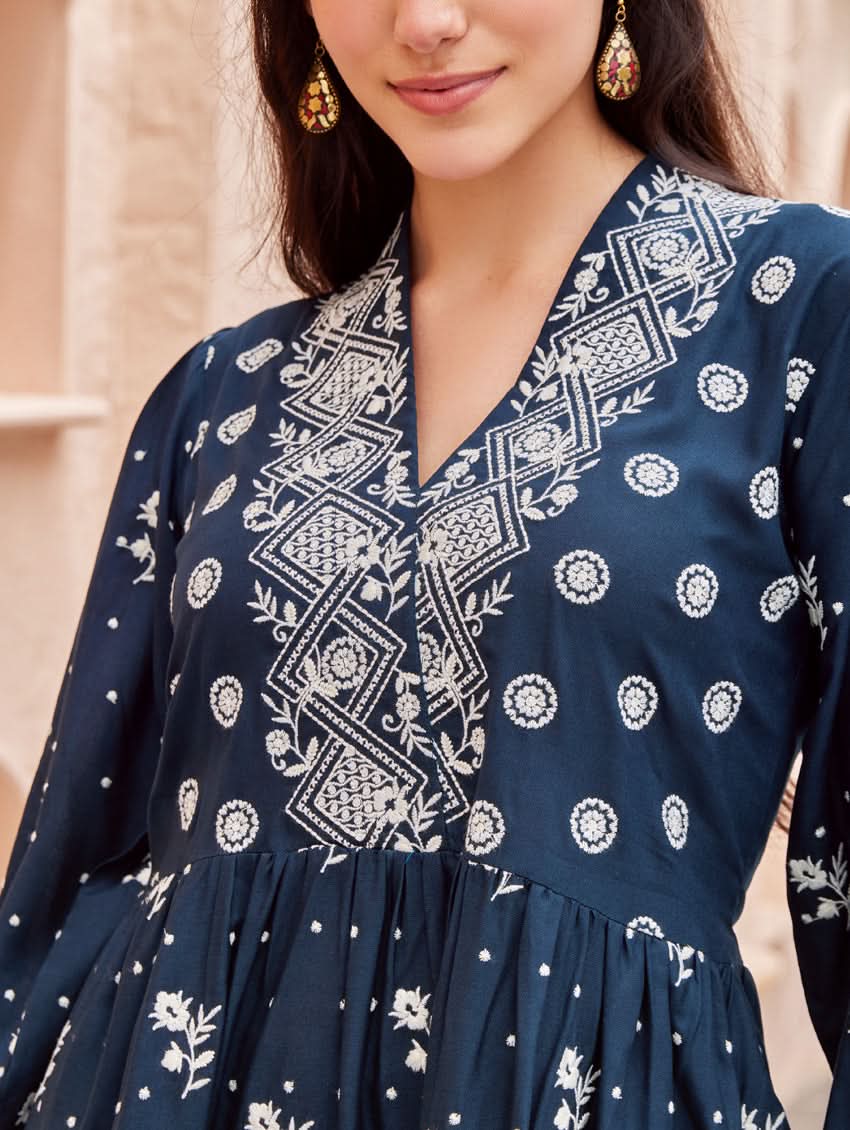 A woman standing on a balcony wearing an embroidered blue kurta set with V-neckline and three-quarter sleeves, paired with black pants with a partially elasticated waistband and pockets on either side.