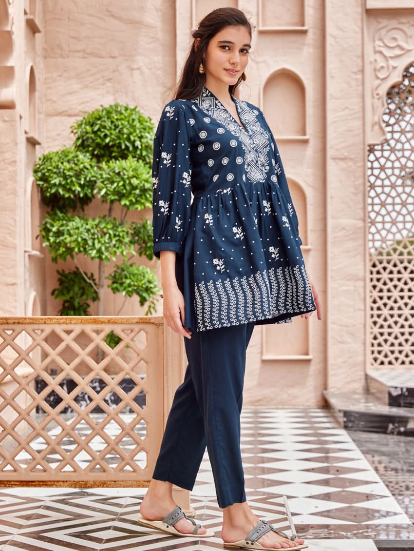 A woman standing on a balcony wearing an embroidered blue kurta set with V-neckline and three-quarter sleeves, paired with black pants with a partially elasticated waistband and pockets on either side.