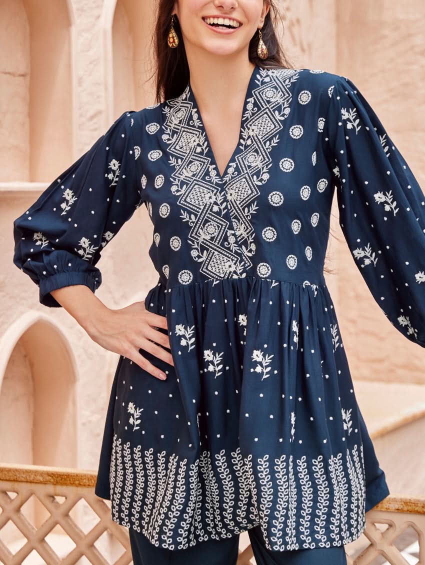 A woman standing on a balcony wearing an embroidered blue kurta set with V-neckline and three-quarter sleeves, paired with black pants with a partially elasticated waistband and pockets on either side.