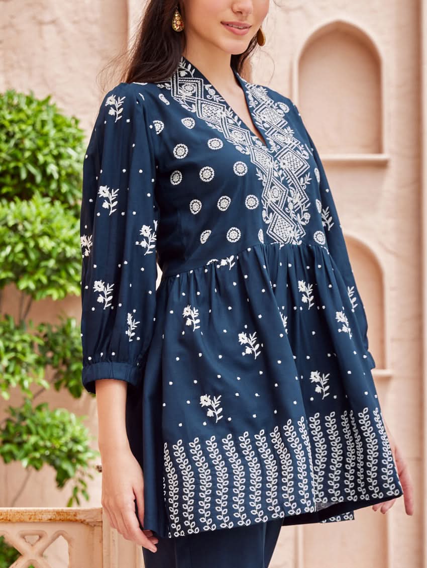 A woman standing on a balcony wearing an embroidered blue kurta set with V-neckline and three-quarter sleeves, paired with black pants with a partially elasticated waistband and pockets on either side.