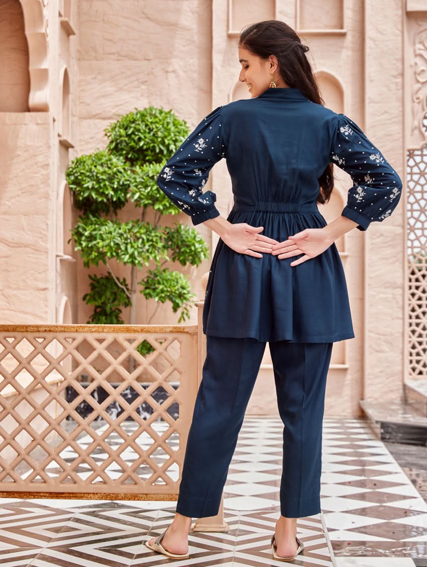 A woman standing on a balcony wearing an embroidered blue kurta set with V-neckline and three-quarter sleeves, paired with black pants with a partially elasticated waistband and pockets on either side.