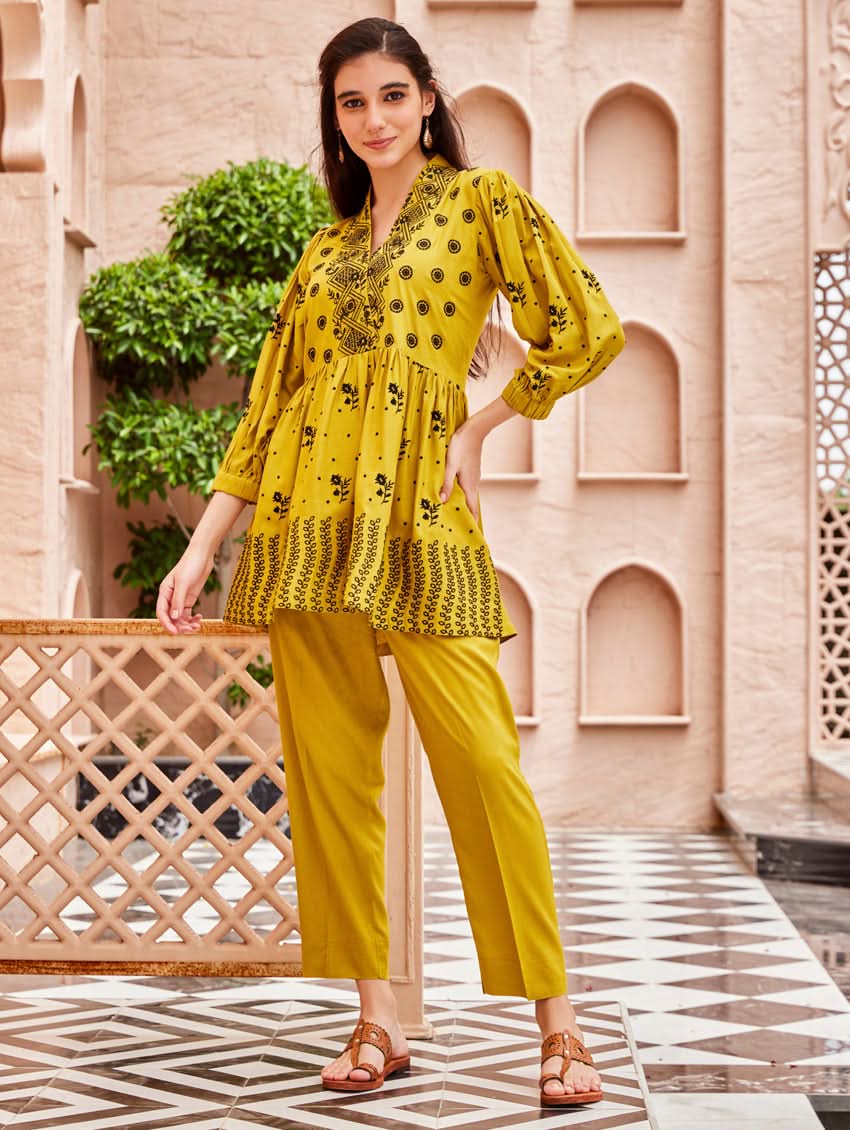 A woman standing on a balcony wearing an embroidered Yellow kurta set with V-neckline and three-quarter sleeves, paired with black pants with a partially elasticated waistband and pockets on either side.