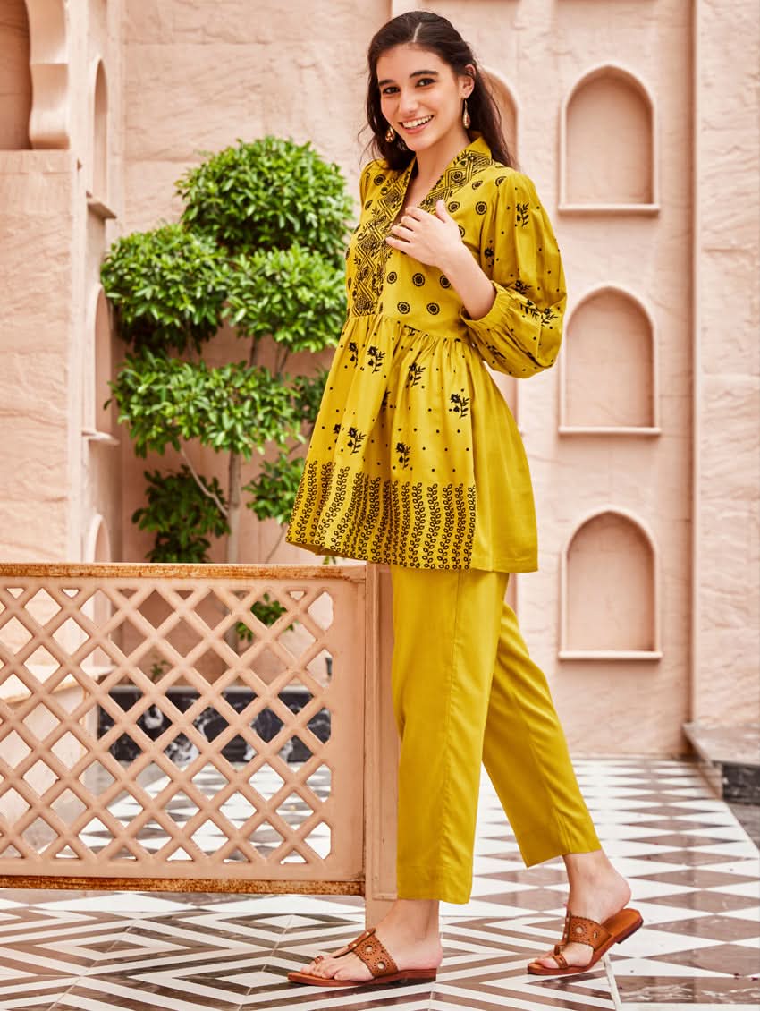 A woman standing on a balcony wearing an embroidered Yellow kurta set with V-neckline and three-quarter sleeves, paired with black pants with a partially elasticated waistband and pockets on either side.