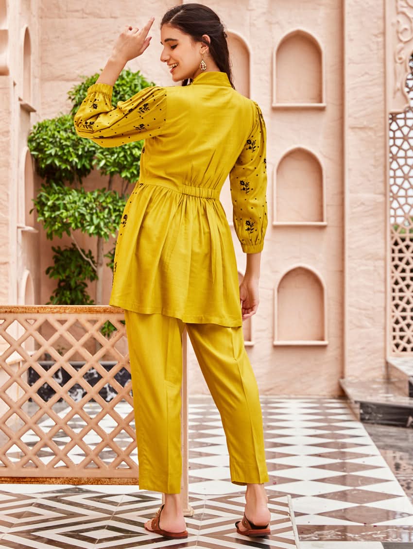 A woman standing on a balcony wearing an embroidered Yellow kurta set with V-neckline and three-quarter sleeves, paired with black pants with a partially elasticated waistband and pockets on either side.