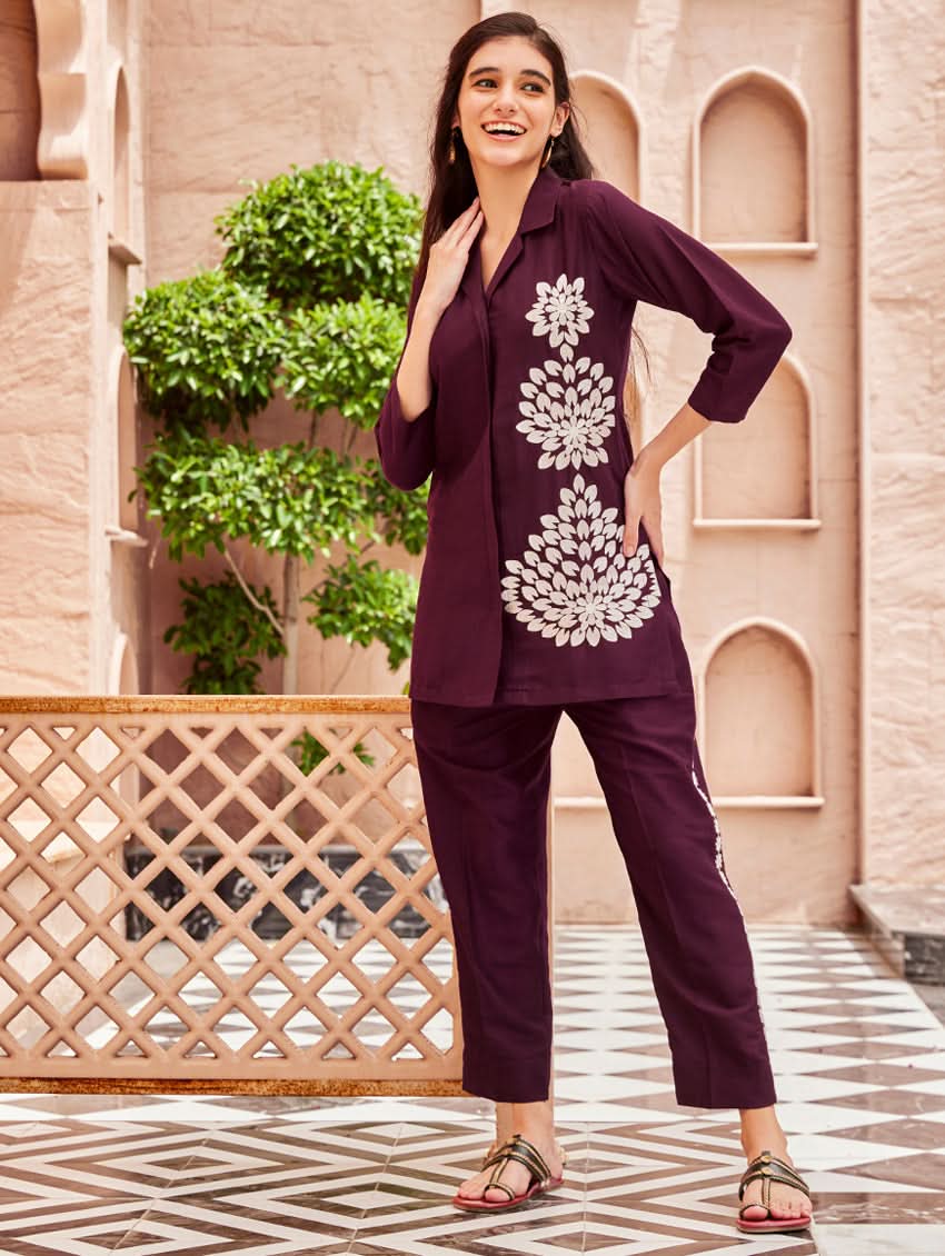 A woman standing outdoors wearing a purple embroidered chunan co-ord set with a revere collar and three-quarter sleeves, paired with solid-colored pants. She is smiling and posing against a backdrop of a pink building with a decorative fence.