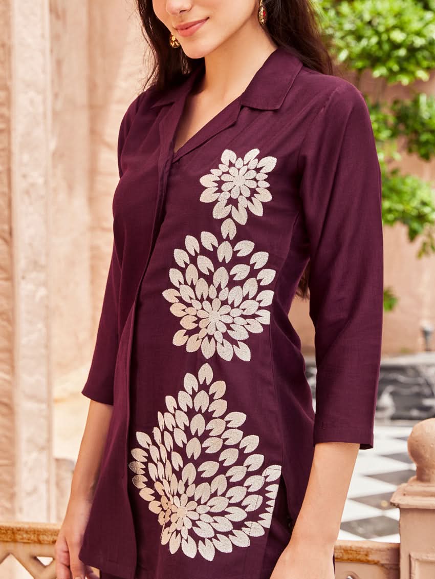 A woman standing outdoors wearing a purple embroidered chunan co-ord set with a revere collar and three-quarter sleeves, paired with solid-colored pants. She is smiling and posing against a backdrop of a pink building with a decorative fence.
