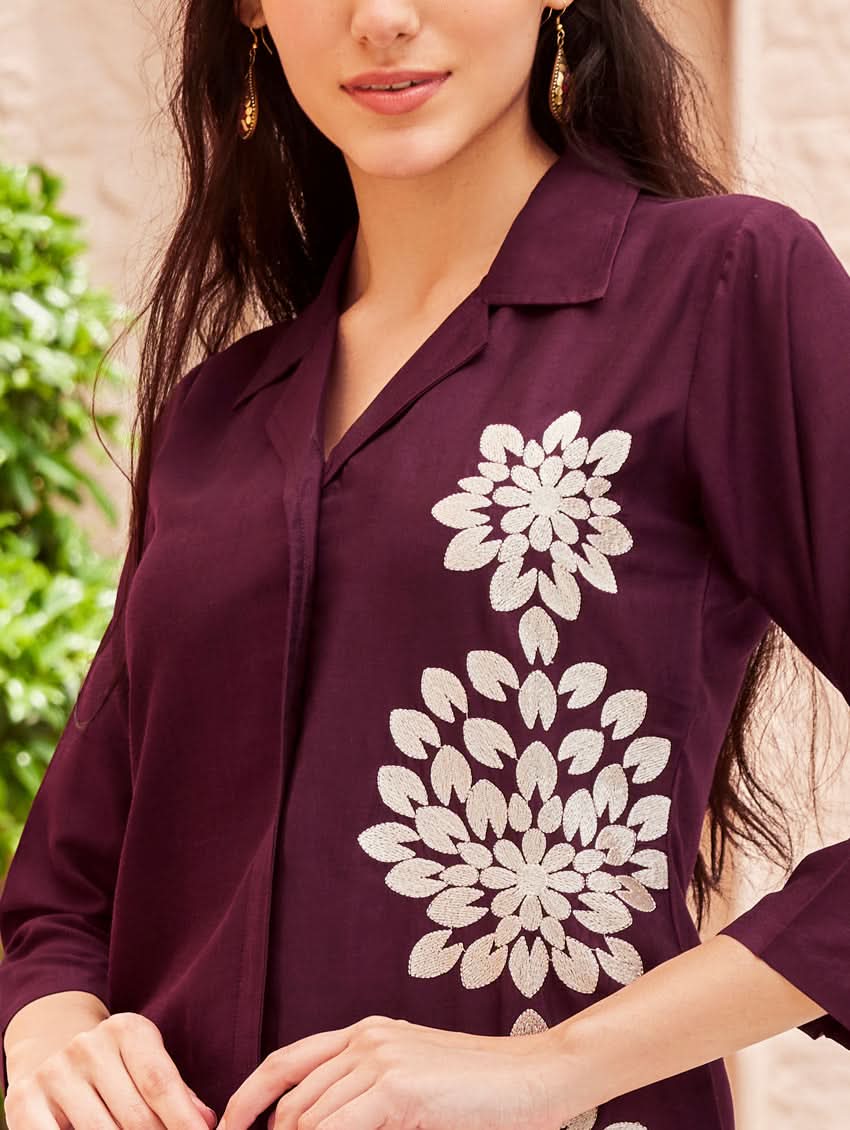 A woman standing outdoors wearing a purple embroidered chunan co-ord set with a revere collar and three-quarter sleeves, paired with solid-colored pants. She is smiling and posing against a backdrop of a pink building with a decorative fence.