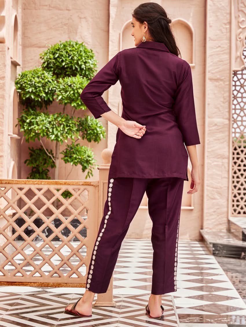 A woman standing outdoors wearing a purple embroidered chunan co-ord set with a revere collar and three-quarter sleeves, paired with solid-colored pants. She is smiling and posing against a backdrop of a pink building with a decorative fence.