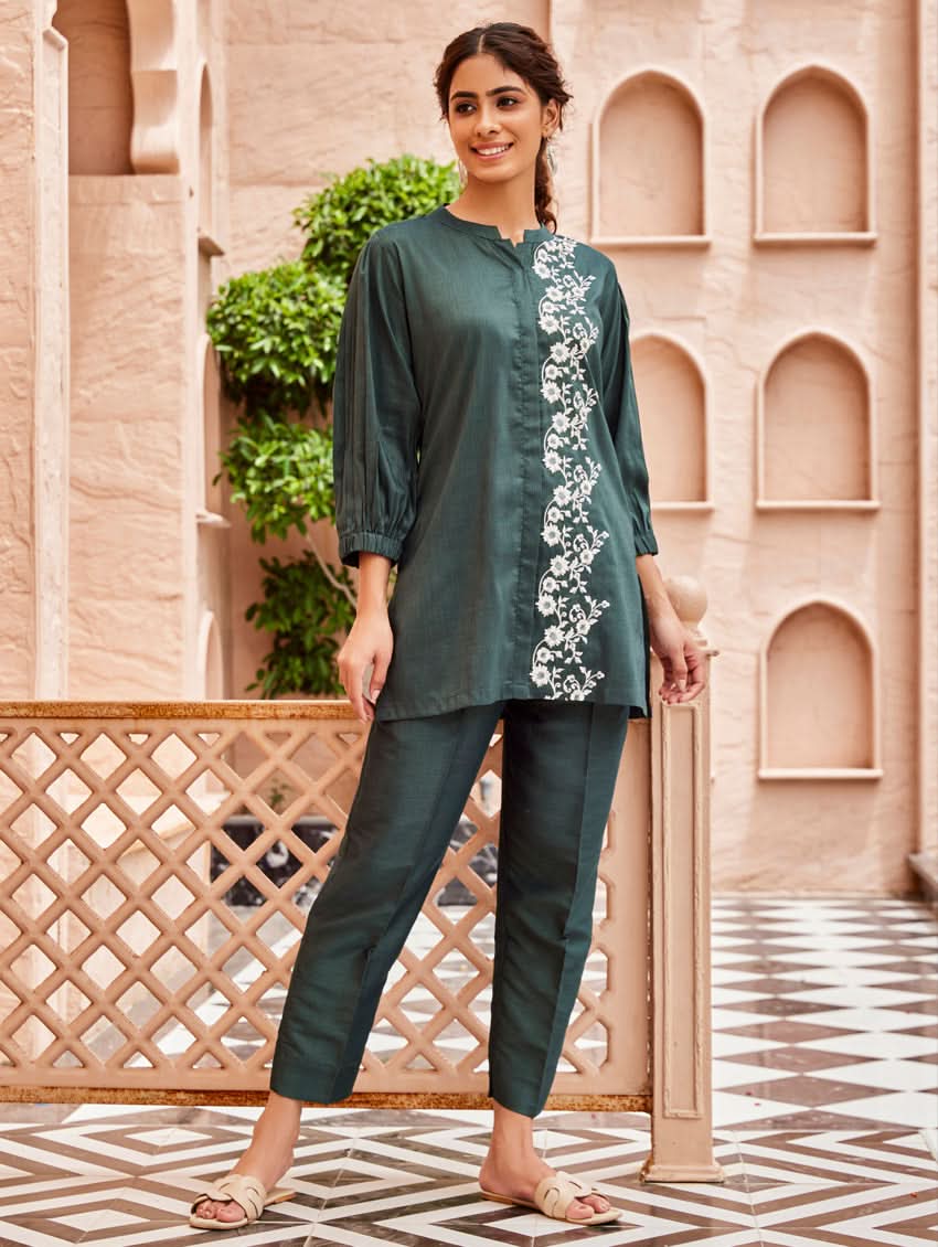 A woman standing in a beautifully decorated area wearing an embroidered green linen silk co-ord set with short sleeves and a partially elasticated waistband. She is also wearing light-colored floral embroidery on the chest area.