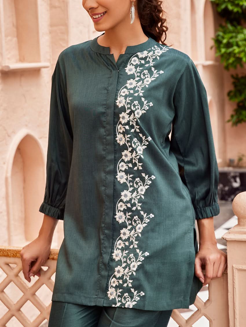 A woman standing in a beautifully decorated area wearing an embroidered green linen silk co-ord set with short sleeves and a partially elasticated waistband. She is also wearing light-colored floral embroidery on the chest area.