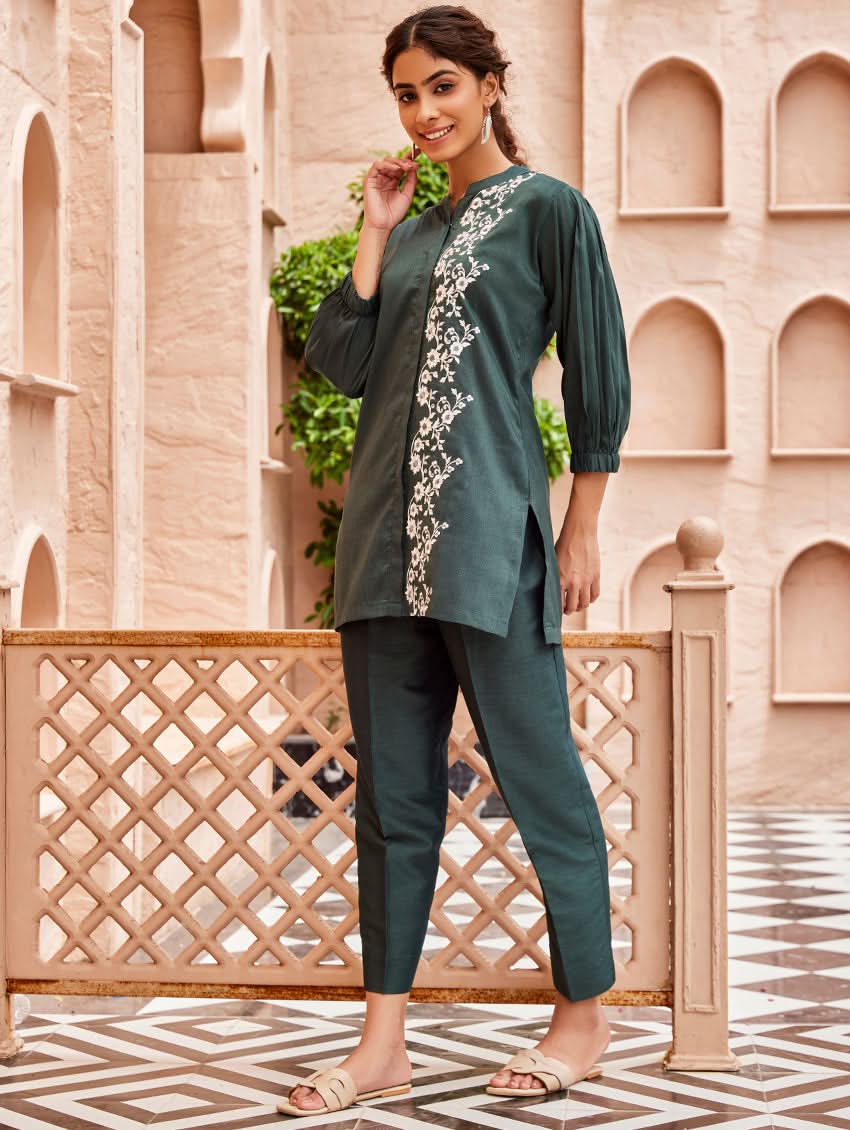 A woman standing in a beautifully decorated area wearing an embroidered green linen silk co-ord set with short sleeves and a partially elasticated waistband. She is also wearing light-colored floral embroidery on the chest area.